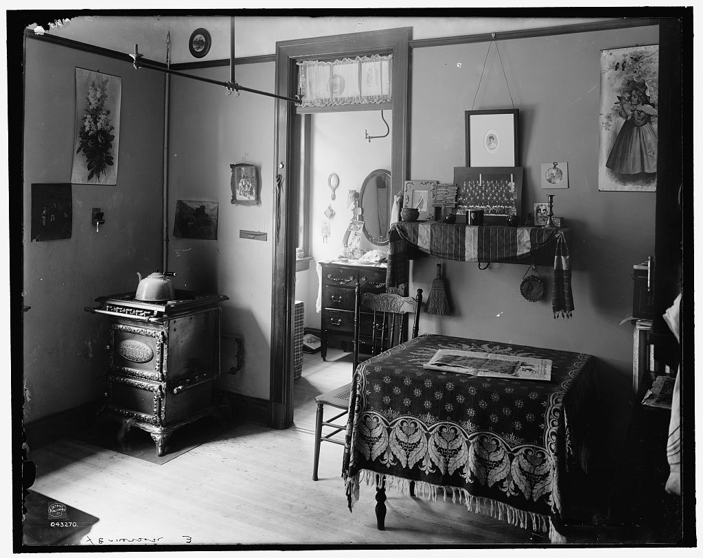 the black and white photograph captures the interior of a simple, early 1900s kitchen.  a woodburning stove sits prominently on the left, with a kettle resting on top.  a wooden table, covered with a patterned tablecloth and a newspaper, occupies the center of the room. a wooden chair is pulled up to the table.  the walls are sparsely decorated with framed pictures and a shelf holds various trinkets. a doorway on the right leads to another room, likely a bedroom, offering a glimpse of a dresser and mirror. the room is bathed in soft, natural light streaming in from the window in the background. the overall impression is one of humble domesticity and a bygone era.