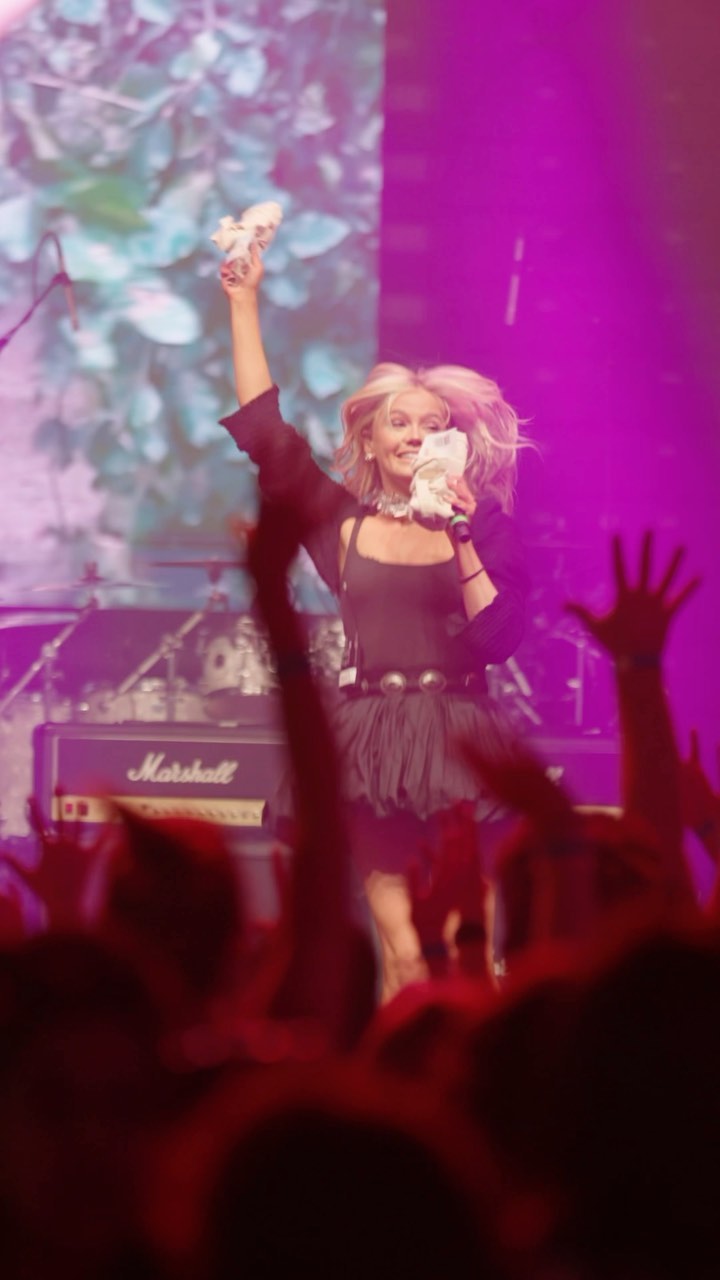 a woman stands on a stage, holding a microphone and money in her hands. she is performing at a concert, with a crowd of people in front of her. behind her is a band and a marshall amplifier. the stage is lit with purple lights.