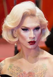 closeup portrait of a blonde drag queen with a vintage hairstyle. she is wearing red lipstick and winged eyeliner, and has floral tattoos on her chest and shoulders. she appears to be at a red carpet event or similar glamorous occasion.  the image focuses on her face and upper body, highlighting her makeup and style.