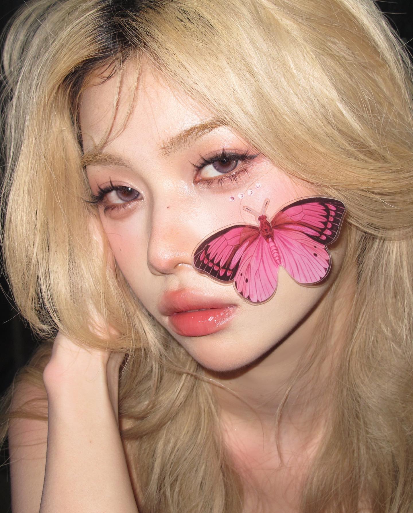 a closeup selfie of a young woman with blonde hair. she has a butterfly sticker on her cheek and glitter makeup under her eye. her makeup look is soft glam with pink tones. she is looking directly at the camera with a neutral expression.