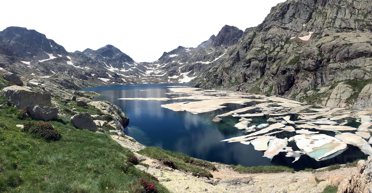 a highangle, wide shot captures a scenic alpine lake surrounded by rugged mountains. the lake is partially covered in ice floes, reflecting the sky and surrounding landscape. green grass and wildflowers grow in the foreground, leading up to the rocky shores of the lake. the mountains in the background are marked with patches of snow, adding to the alpine setting.