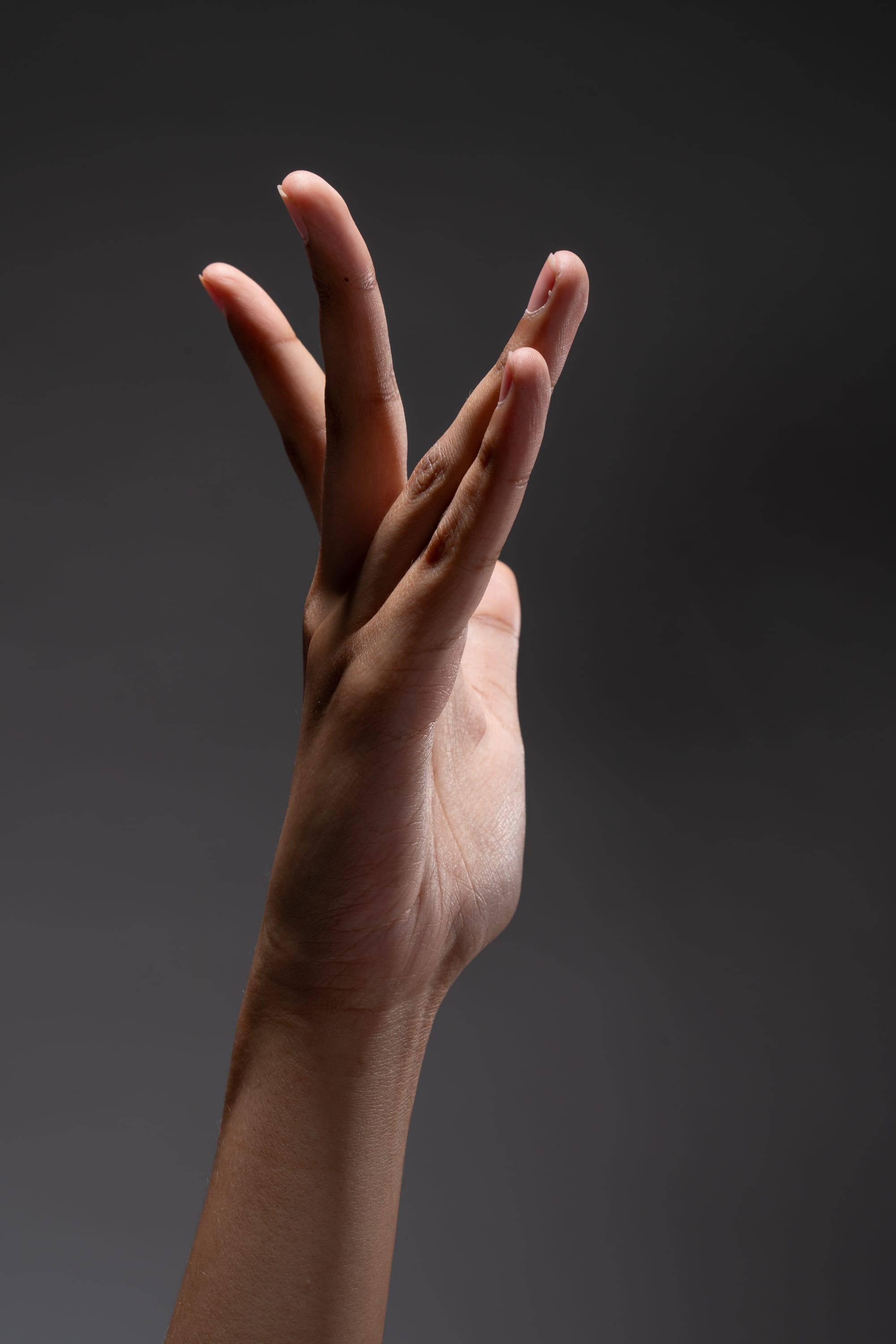 a hand is extended against a dark gray background. the fingers are curled in a unique gesture. the image is illuminated with dramatic lighting, casting shadows on the hand and creating a sense of depth.