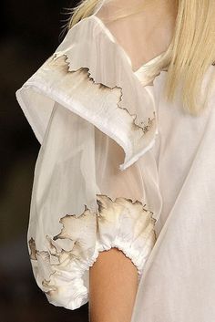 a closeup shot of a model wearing a white sheer dress with delicate ruffles. the dress is flowing and ethereal, with a soft, feminine quality. the model's arm is visible, showcasing the intricate details of the sleeves.  the fabric is luxurious and textured, adding to the overall elegance of the piece. the dress is a perfect example of high fashion and couture.
