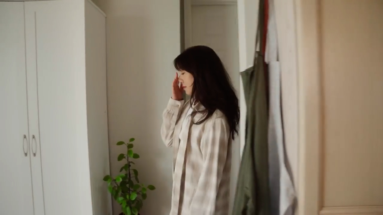a young woman with long, dark hair is standing in a hallway. she is wearing a beige and white plaid shirt. she is rubbing her eye with her right hand, as if she is tired or just waking up. the walls and doors in the hallway are white. there is a green plant in a pot on the left side of the image. the image is lit by natural light coming from the left side of the frame.