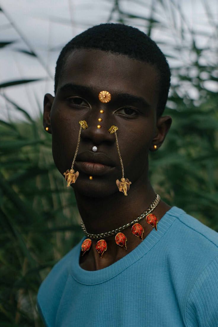 a portrait of a darkskinned man with multiple pieces of ornate jewelry. he has a circular gold piece on his forehead, several small gold dots running down his face, a barbell nose piercing, and intricate gold nose jewelry with small cherub figures hanging on either side of his mouth. he also wears gold earrings and a chain necklace with alternating red and gold petalshaped charms. the man is wearing a light blue crewneck shirt, and the background consists of blurred green foliage.