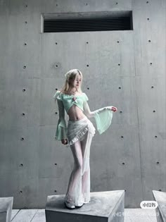 a woman is posing for a photo shoot. she is wearing a white skirt, a light green top and white heels. she is in an indoor studio with a white wall and grey floor. the woman is looking at the camera and she is smiling.