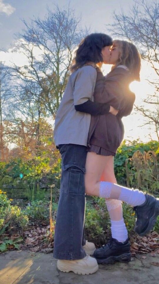 a young couple is standing in a forest and hugging each other. the woman is wearing a brown sweater and a black skirt and her boyfriend is wearing a white shirt and blue jeans. they look very happy and in love with each other. they are kissing as the sun beams down on them.  they are wearing stylish footwear and white socks with black accents.