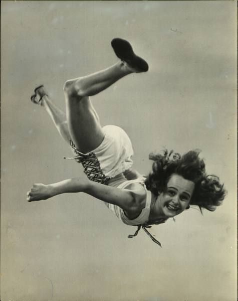 a young woman in a tank top and shorts jumps through the air during a high jump competition. the black and white image captures her midair with a joyful expression on her face. her blonde hair flows behind her.  the image was captured in a vintage outdoor setting.