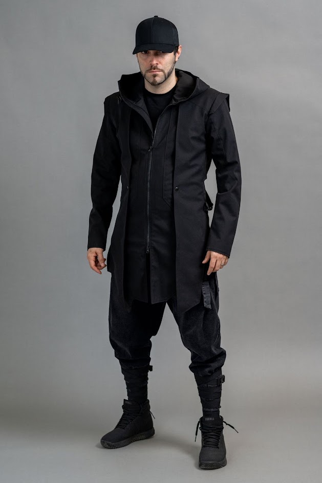 a man wearing a black coat with a hood, black pants, black boots, and a black cap is standing in front of a grey background, looking directly at the camera.