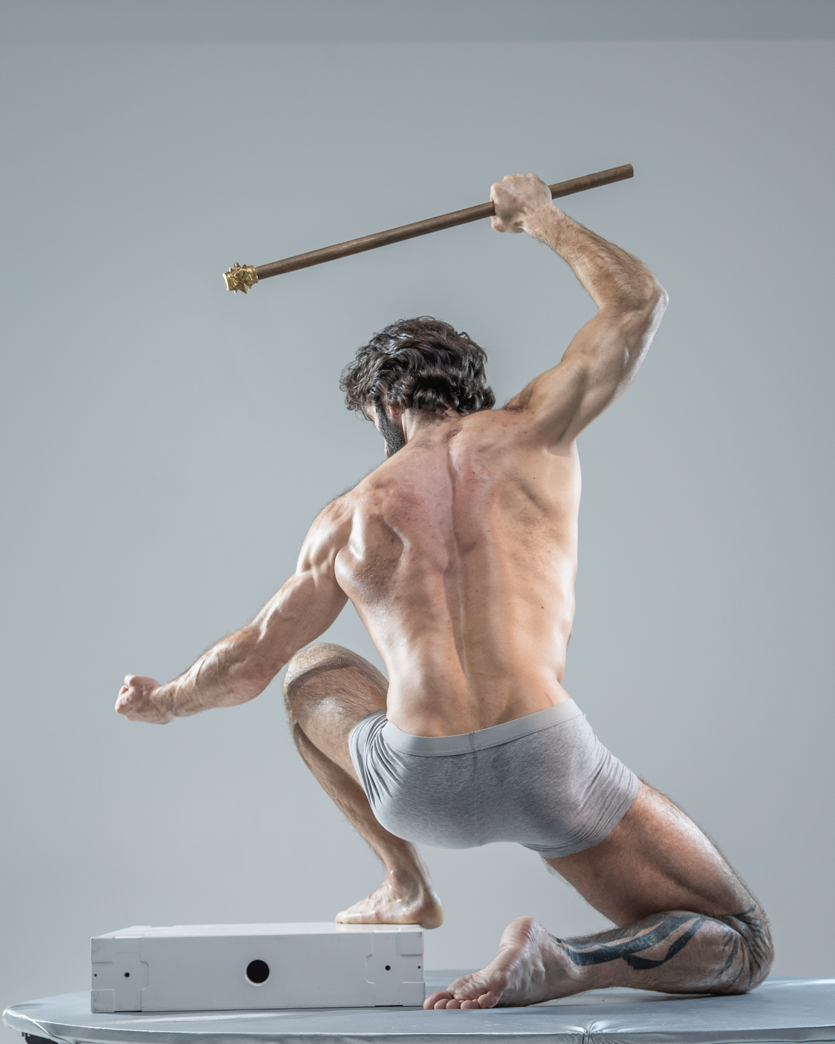 a muscular man with long hair and a beard crouches on the floor, poised for action. he holds a wooden staff above his head, his body tensed with energy. he wears only grey underwear, his physique on full display against the plain studio background.  the image evokes a sense of power and dynamism, captured with strong lighting that highlights the contours of his muscles.