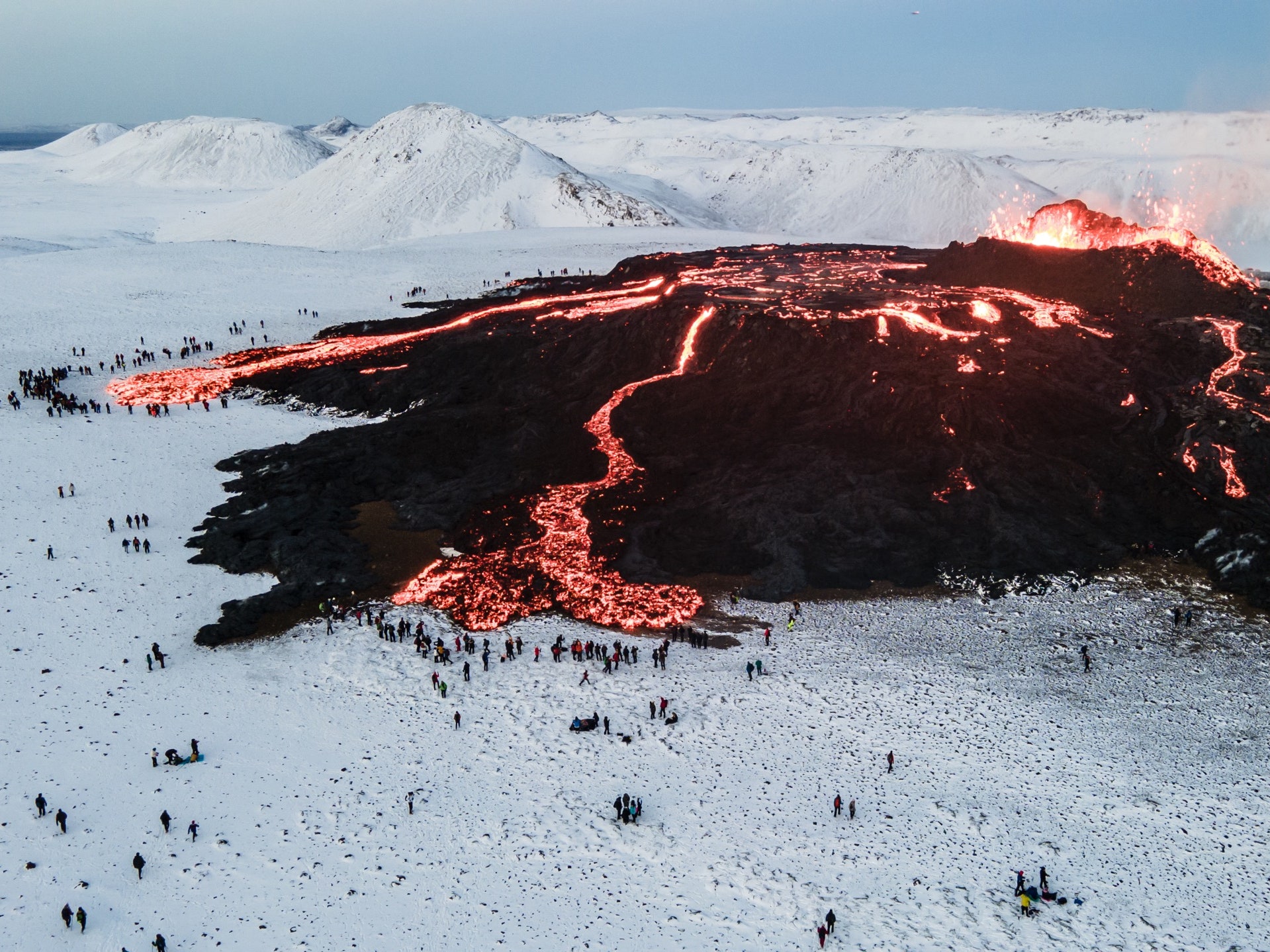 an aerial view of a volcano erupting with lava flowing down the mountainside into a snowcovered landscape. the image shows a stark contrast between the fiery red lava and the pristine white snow. people are gathered on the surrounding snow, observing the spectacle.