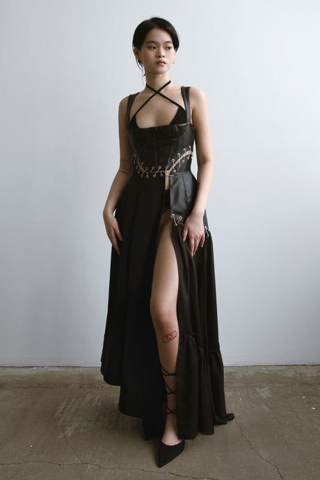 a fulllength shot of a fashion model posing in a stylish black dress. the dress features a leather corset top with laceup detailing and a flowing maxi skirt with a high leg slit. the model is wearing black laceup high heels and has a minimalist look with her hair pulled back. the background is a simple, neutral grey. the overall style is dark, elegant, and fashionforward.