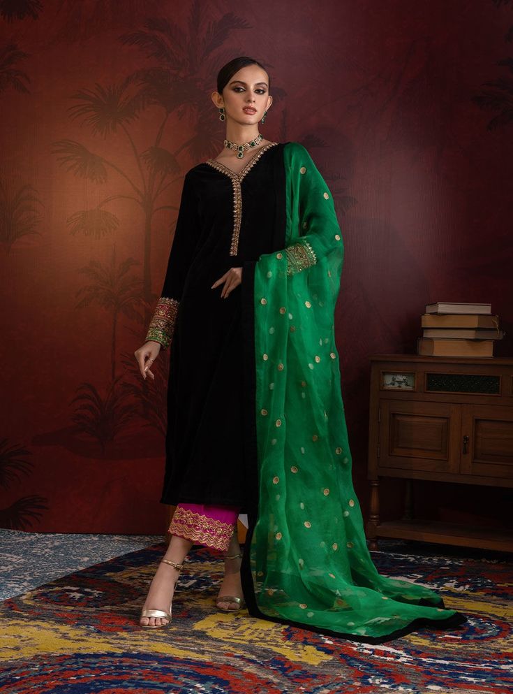 a woman is standing and posing in a black velvet shalwar kameez with a vibrant green dupatta. the shalwar kameez is adorned with delicate gold embroidery, and the shalwar features a pink border.  she's wearing gold jewelry. the green dupatta has gold accents and is elegantly draped around her.  the background is a rich maroon with a palm tree print. the setting includes an ornate area rug and dark wood furniture. the overall aesthetic suggests a highend fashion editorial shoot showcasing traditional south asian attire.