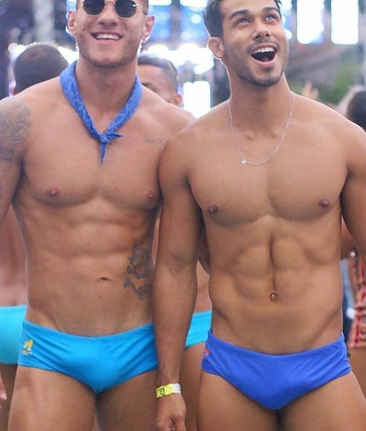 two men, both shirtless and wearing blue swim trunks, stand side by side and smile for the camera. the man on the left has a blue bandana tied around his neck, while the man on the right has a silver necklace. they are both muscular and appear to be having a great time in the sun.  the photo is vibrant and joyful, and celebrates the beauty of diversity.