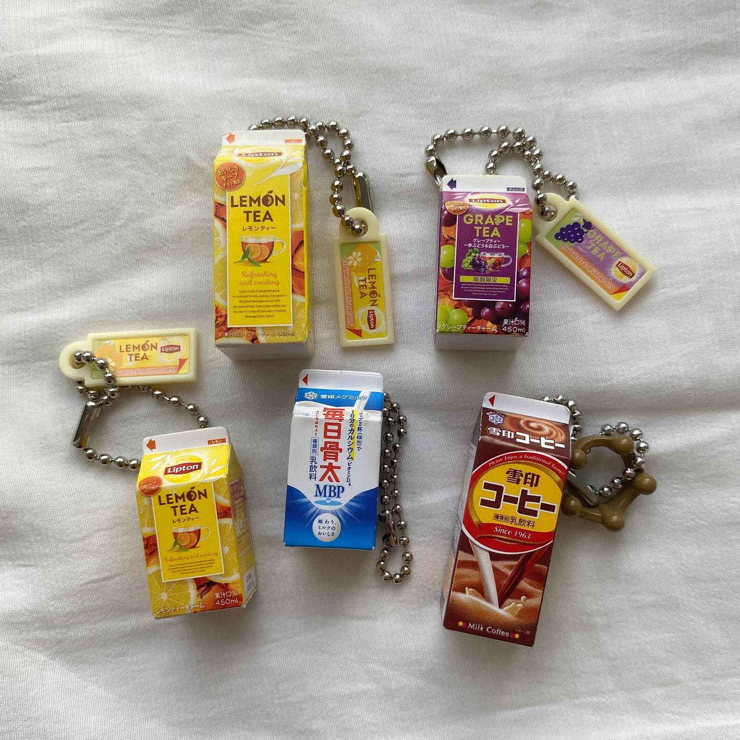 the image shows a collection of miniature beverage carton keychains displayed on a textured white surface. there are two lipton lemon tea keychains, a grape tea keychain, a milk coffee keychain, and a "mainichi honebuto" milk keychain, all attached to chains.