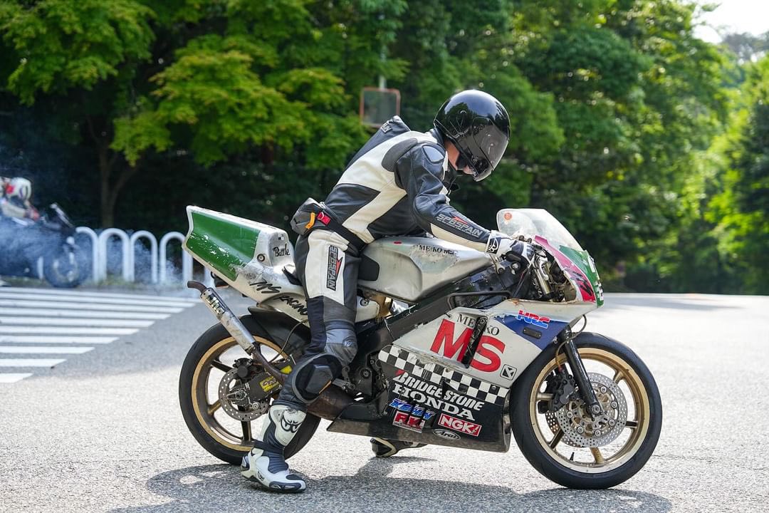 a motorcyclist in full leathers and a helmet is leaning into a corner on a racetrack. the motorcycle is a honda, with a number plate and sponsor logos. the rider is wearing a white and black racing suit, and the motorcycle is white with green and red accents. the background is a green forest. the sun is shining brightly, and the asphalt is dry.  the photo captures the intensity of the race, and the skill of the rider.