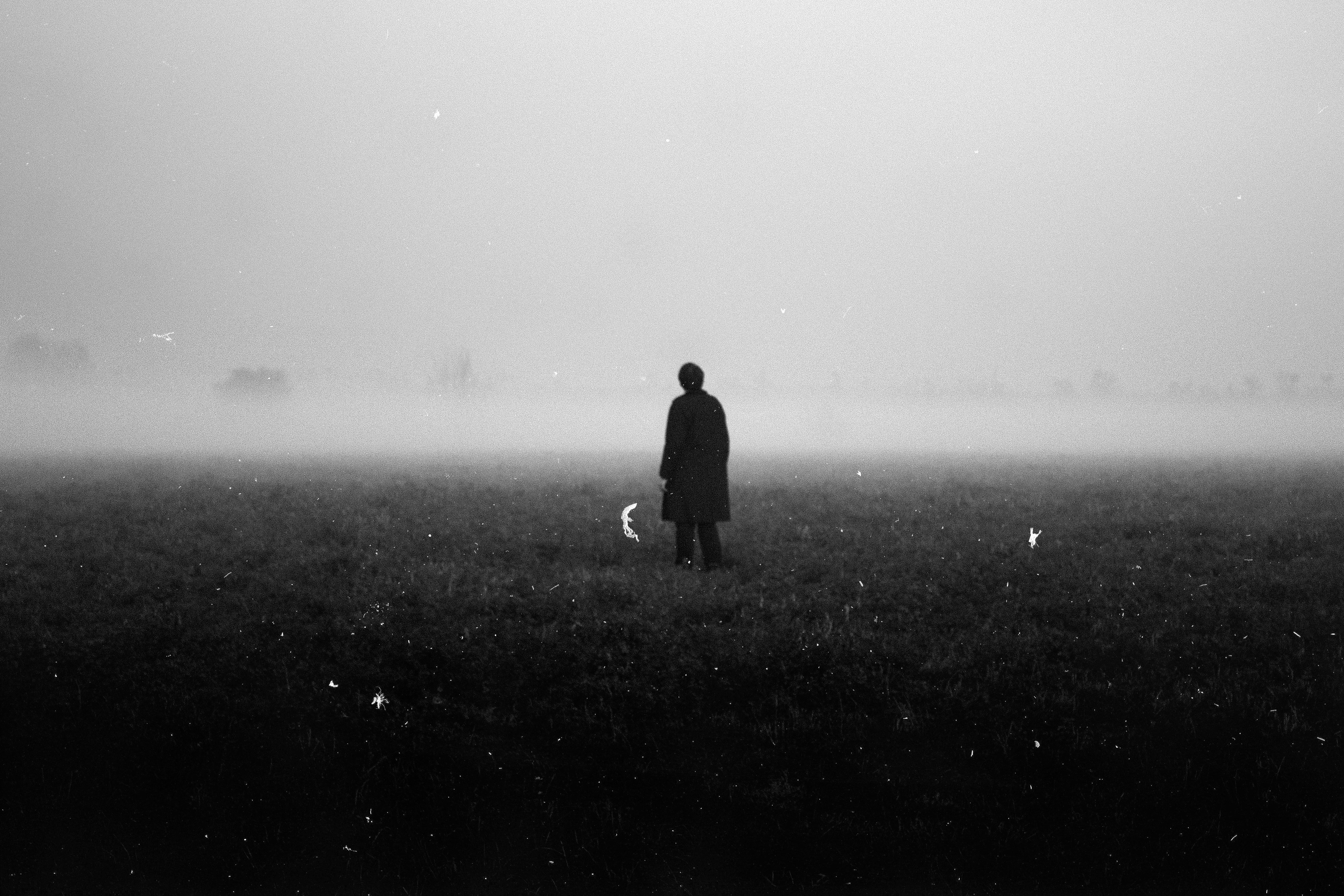 a lone man stands in the center of a foggy field. he is facing away from the camera, his figure silhouetted against the dense mist. the grass is short and covered in dew, and the fog obscures the horizon, creating a sense of isolation and vastness. the image is rendered in black and white, adding to its stark beauty and evoking a feeling of mystery and contemplation.
