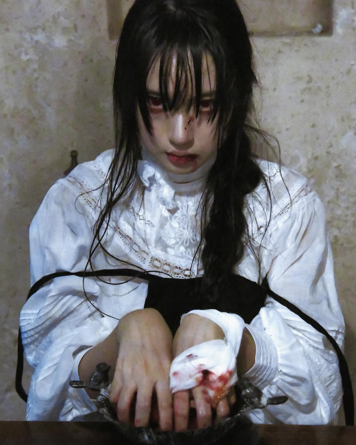 a woman with dark, wet hair and pale skin, exhibiting red eyes, is depicted in an eerie, gothic setting. she has a bloody injury on her face and blood on her lips. a white blouse with lace details and a black bodice adorns her. she holds a bowl with blood, in her hands one of which is wrapped in bandages. the antique or vintage aesthetic adds to the overall scary and dramatic atmosphere of the indoor portrait.