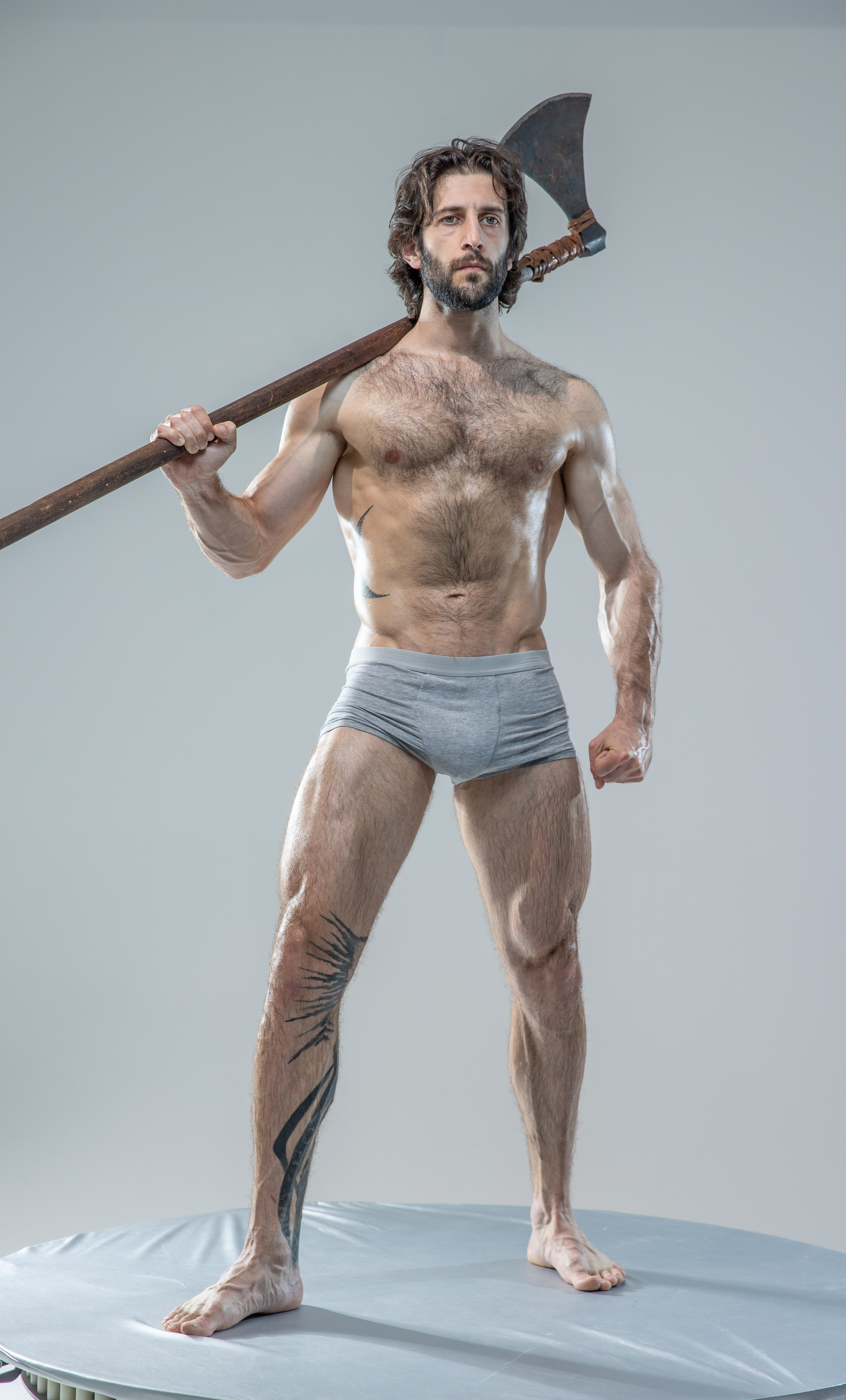a muscular, shirtless man with long hair and beard stands on a round platform in a studio setting. he holds a large, battleworn axe over his shoulder, his expression serious. he's wearing only grey underwear, showcasing his toned physique and a tattoo on his left leg. the lighting is bright and artificial, highlighting his muscular build.