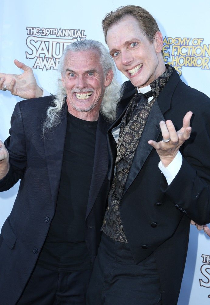 two men are smiling and posing for a picture while standing next to each other at a red carpet event. they are wearing black suits with the man on the right wearing a tuxedo. the man on the left is wearing a black jacket over a black tshirt and the man on the right is wearing a white shirt with a black patterned tie. the background is a white wall with the logo of the awards show and a blue wall to the left.