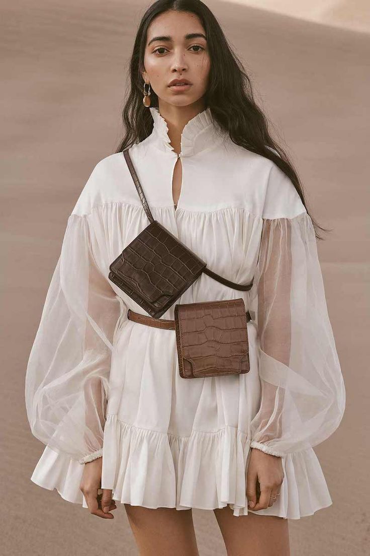 a young woman with long dark hair is modeling a white mini dress with voluminous puff sleeves and ruffles. the dress has a high neck and is made of a lightweight, slightly sheer fabric. she is wearing a brown crocodileembossed leather crossbody bag with two separate compartments, and gold hoop earrings.  the setting appears to be a sandy desert landscape. the overall style is elegant and summery.