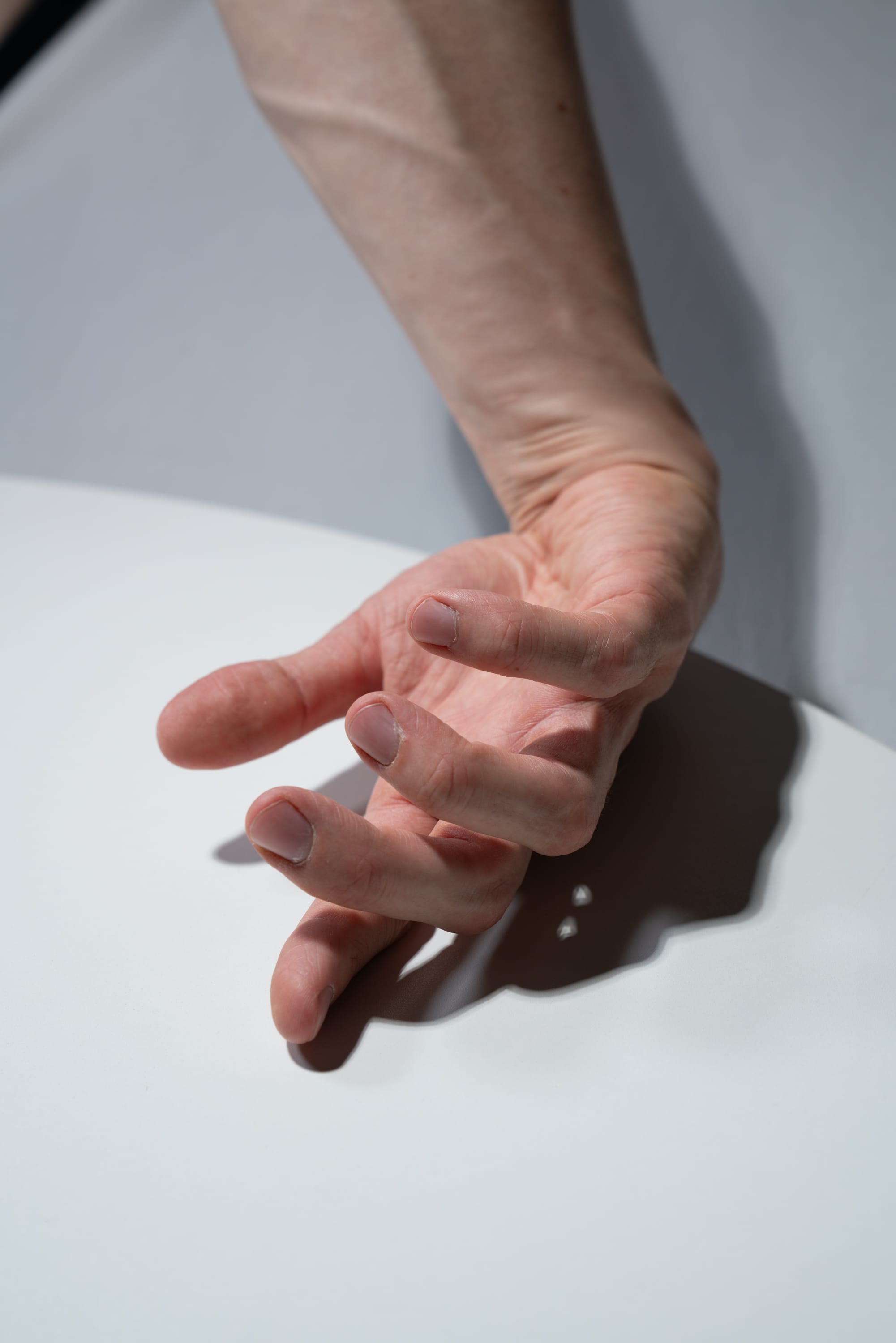 a closeup image of a man's arm and hand resting on a white table. the hard light casts a distinct shadow of the hand on the table's surface. the image focuses on the details of the hand, including the veins and wrinkles, with a minimalist aesthetic.