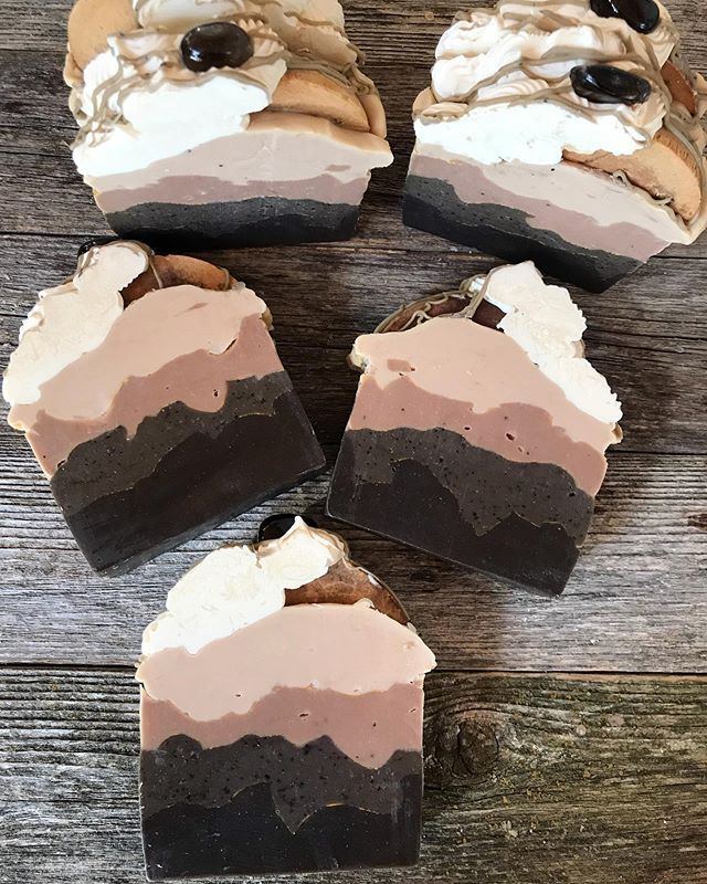 a close up of five bars of soap, all made to look like coffee and whipped cream desserts. the soaps are all a mix of brown and white, with the brown being the coffee part and the white being the whipped cream. they have a layered look, and there is a hint of chocolate on some of the bars. the soap is on a rustic wooden background, giving the image a natural and handcrafted feel.