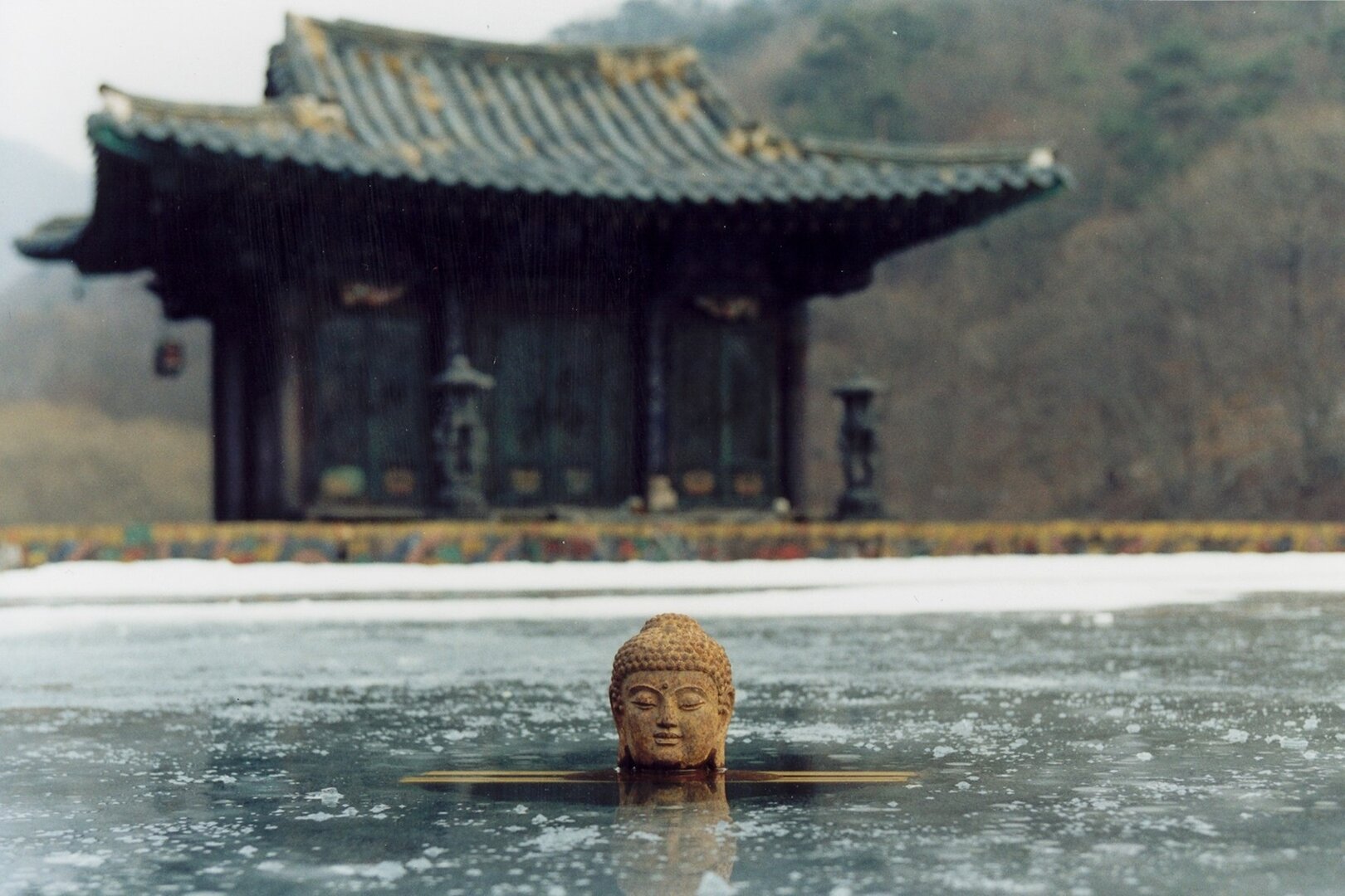 a serene winter scene captures a buddha statue's head partially submerged in a frozen pond. the icecovered water reflects the overcast sky, creating a muted palette of grays and blues.  in the background, a traditional east asian temple stands majestically, its dark wooden structure dusted with snow. the composition evokes a sense of tranquility and spiritual reflection.