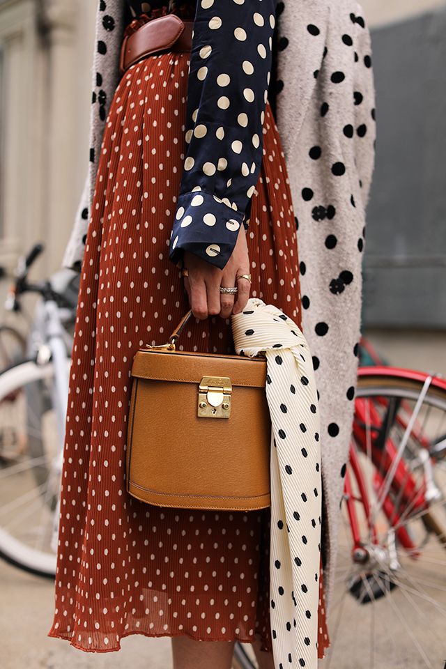 a woman is standing outdoors wearing a rustcolored polka dot midi skirt, a navy polka dot shirt, and a grey cardigan. she is carrying a brown boxshaped handbag and a creamcolored polka dot scarf. the photo is taken from the waist up and shows the woman's outfit and accessories in detail. the background includes some parked bicycles, suggesting an urban setting.