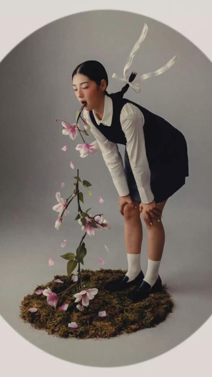 a young asian woman in a black dress and white shirt is elegantly bent over a small plant with pink magnolia flowers.  she appears to be delicately eating or interacting with one of the blossoms. the scene is set against a neutral gray background, with the plant nestled in a patch of moss. the overall aesthetic is artistic and slightly surreal, with a fashionforward feel. the girl’s ponytail is adorned with a white ribbon.