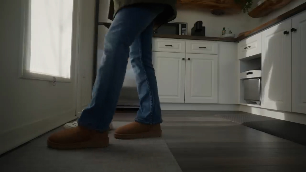 a woman wearing blue jeans and brown ugg boots is walking towards the camera from a doorway into a kitchen. the kitchen has white cabinets, dark wood floors, and stainless steel appliances. the image is taken from a low perspective and the lighting is bright and natural.