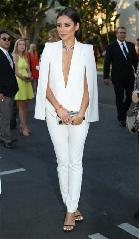 a stylish woman is captured in a full shot, elegantly dressed in a white pantsuit featuring a capelike blazer. the pantsuit is impeccably tailored, accentuating her figure.  she pairs the ensemble with simple black heeled sandals and a striking necklace. the setting appears to be an outdoor event, possibly a red carpet, as evidenced by the presence of other welldressed individuals in the background, including men in suits. the overall impression is one of sophistication and high fashion.