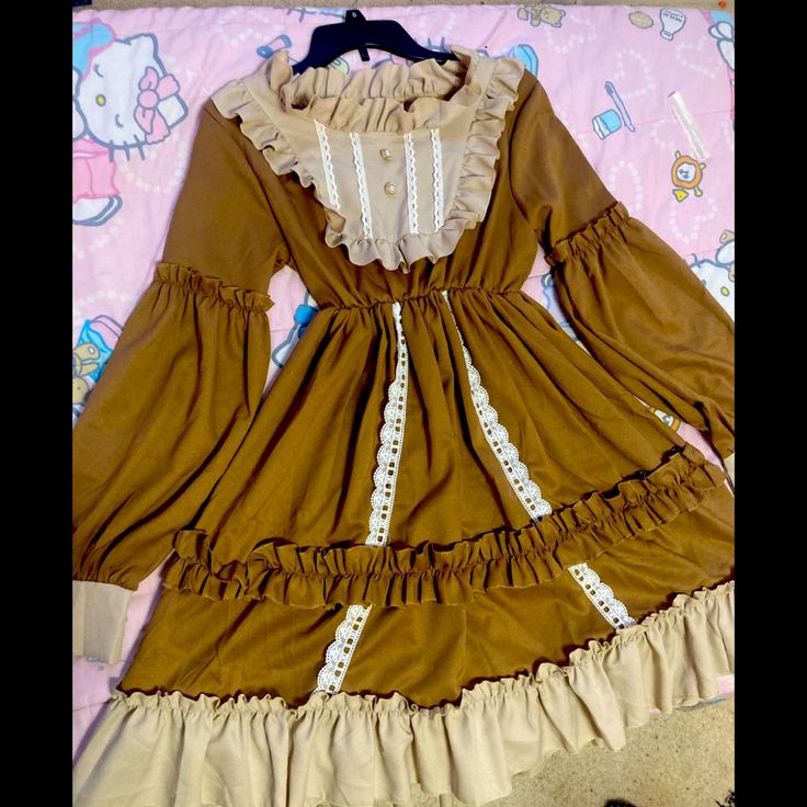 a brown and cream lolita fashion dress laying on a bed. the dress has long sleeves, a ruffled collar and skirt, and lace accents. the background shows a bed with a colorful blanket.