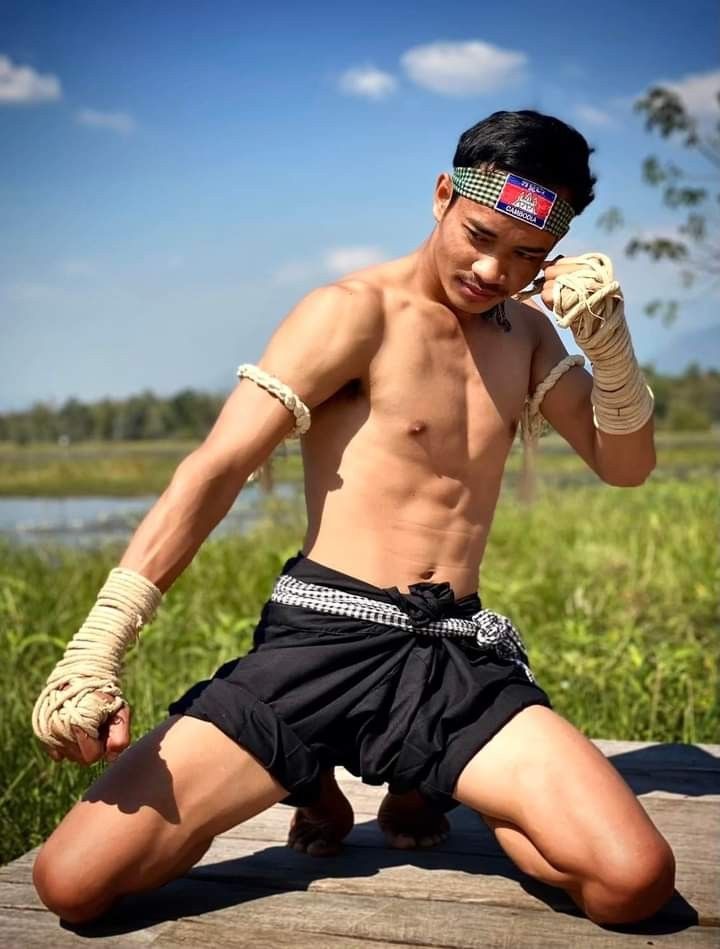 a young man, shirtless and wearing traditional muay thai shorts, kneels in a fighting stance with his hands wrapped in bandages. he is muscular and has a sixpack abdomen. he is looking at the camera with a focused expression. the background is a grassy area with a wooden walkway and a blue sky.