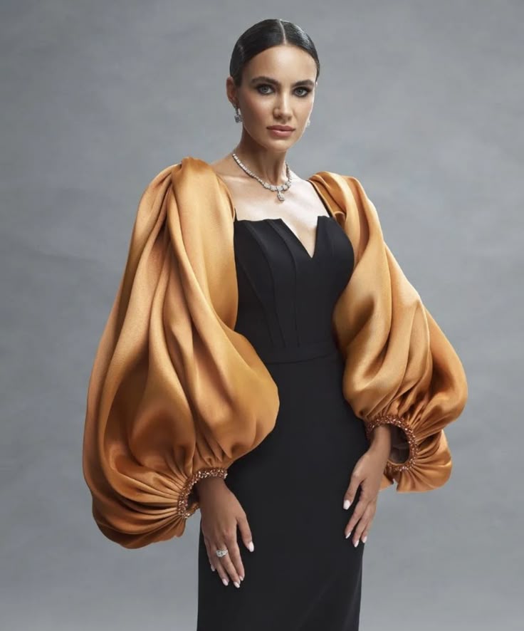 a woman in a black dress with dramatic gold satin puff sleeves stands in front of a grey background. she is wearing elegant jewelry including a necklace, earrings, and a ring. her hair is styled in an updo, and she is wearing makeup. she has a serious expression on her face.