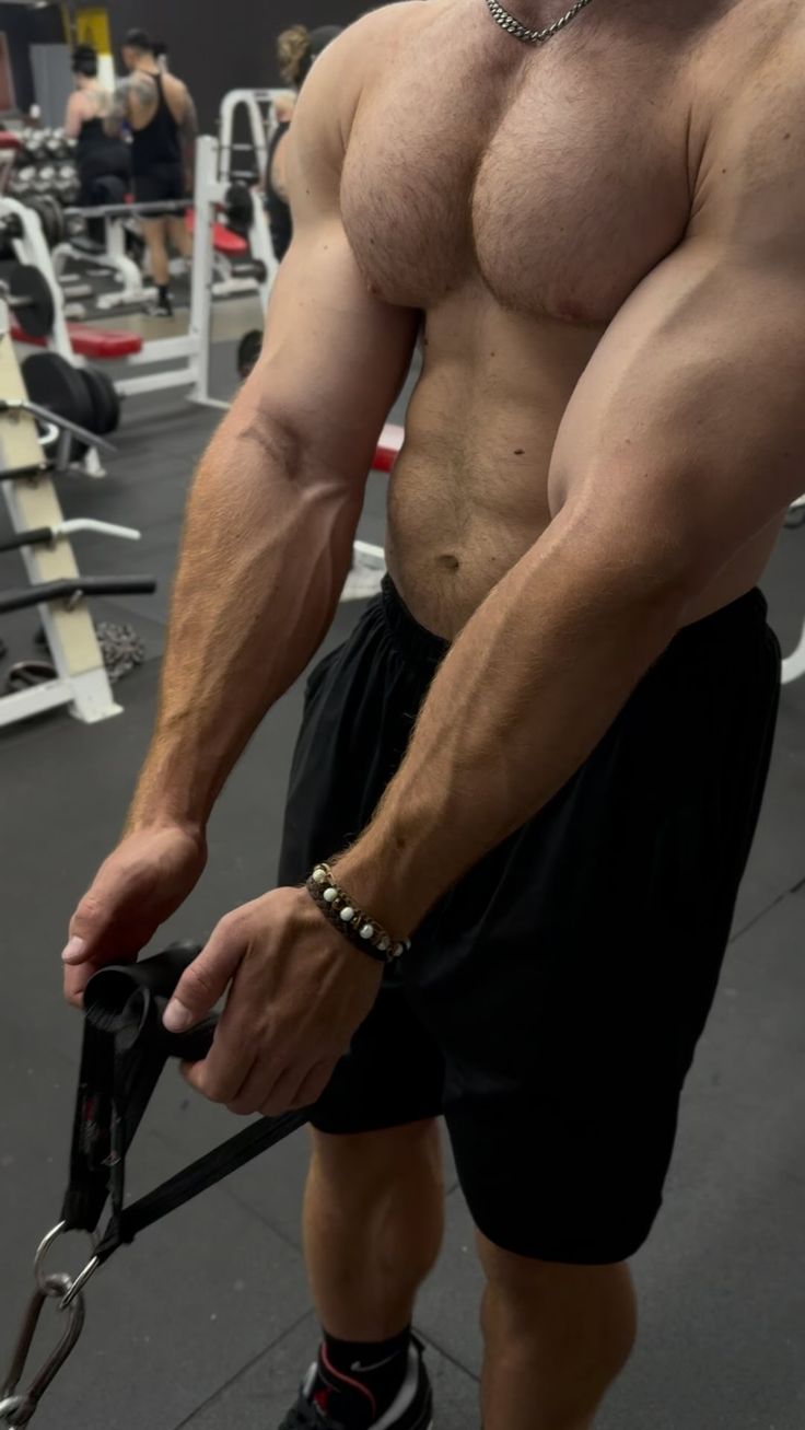 a shirtless man with a muscular physique is working out at the gym. he is using a cable machine and pulling on a cable attached to a handle with both hands. he is wearing black shorts and has veins visible in his arms. the man has a strong chest, defined abs, and muscular biceps and triceps. he is focused on his exercise and is sweating slightly.  the image captures the intensity and dedication of a serious weightlifter.