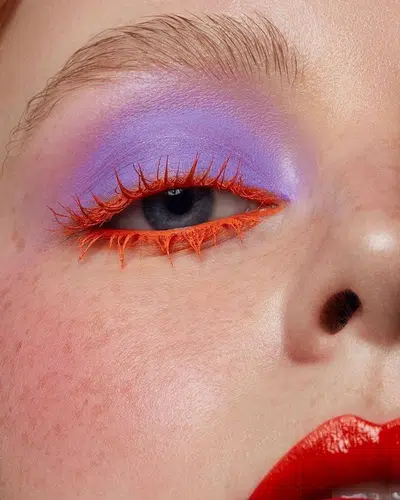 a close up of a woman's face with her eye makeup done in purple and orange. she has on a red lipstick and has a beauty mark on her cheek.  the background is blurry.