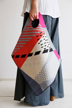 a woman wearing gray pants is holding a colorful bag with a geometric design in a variety of pink, red, white and gray colors. the bag is a mix of different patterns, including stripes, triangles, and other shapes. the woman is standing and is mostly out of frame.