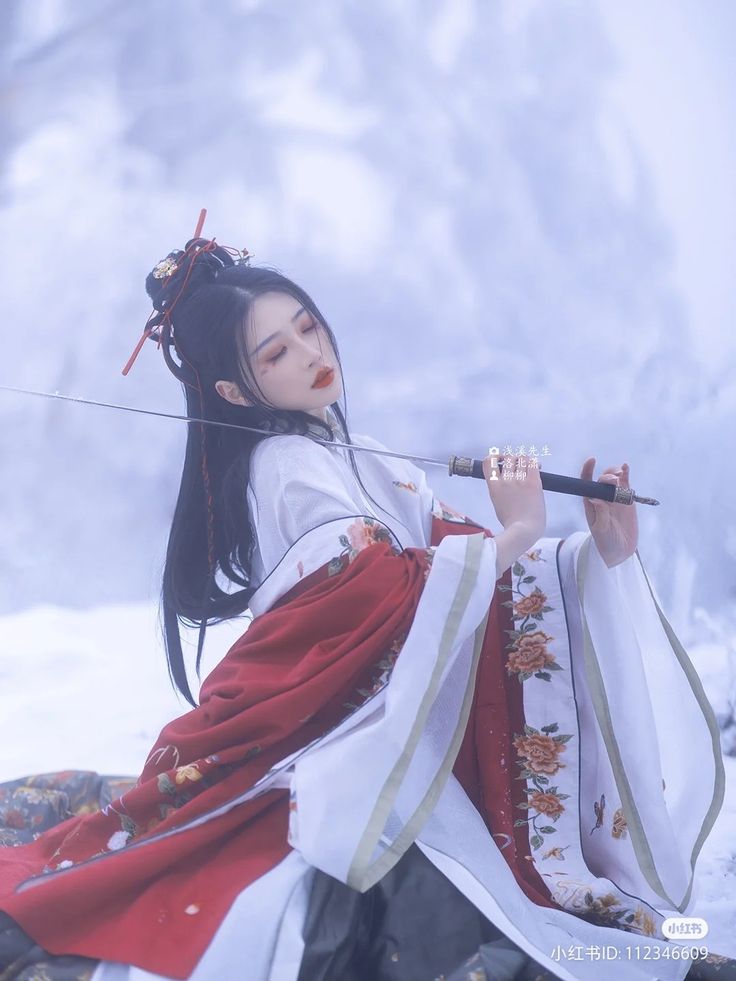 a beautiful young woman in a traditional chinese dress, holding a sword, stands in a snowy winter landscape. she is dressed in a red and white robe, with her long black hair flowing behind her. the background is a soft blur of white snow and blue sky.