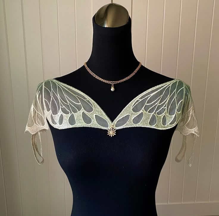 a black mannequin is shown with a gold chain necklace, a white pearl drop pendant, and a fairywingshaped shoulder cover with green lace and a sparkling rhinestone snowflake embellishment in the center. the background is a vertically paneled beige wall.