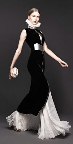 a woman stands in a black and white dress against a gray background. the dress has a ruffled collar and is fitted to her waist. the skirt is a white flowing fabric and is gathered at the bottom.  she is wearing a silver belt with a small purse in her left hand.  she has her left hand on her hip, with her right hand extended at her side.  she is looking off to the side with a serious expression.