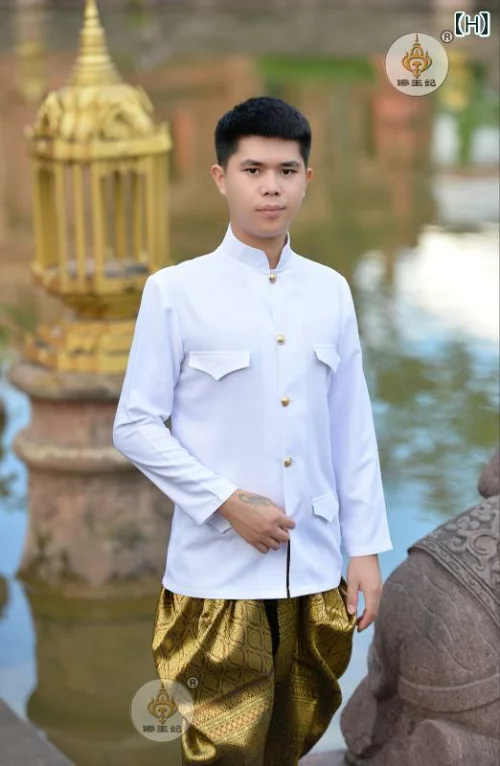 a young thai man is wearing a traditional white shirt and gold pants, standing in a garden. he is smiling and looking at the camera. there is a statue of a lion behind him. there is a pool of water reflecting sunlight.