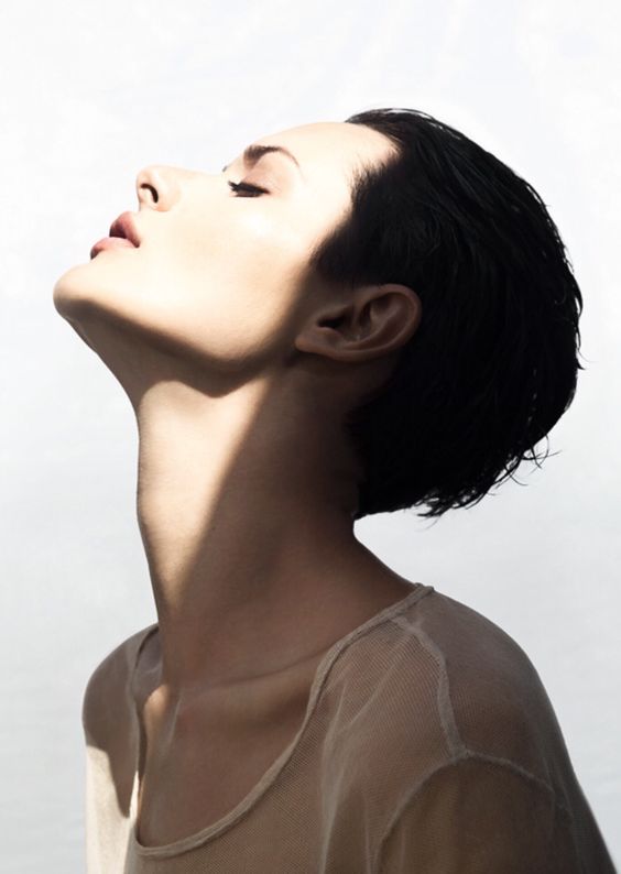 a woman with short black hair is shown in profile with her head tilted back, her eyes closed, and her face bathed in sunlight. the hard light creates strong shadows on her neck and face, emphasizing her bone structure and the delicate lines of her profile. the woman's simple, sheer, beige top adds to the minimalist aesthetic of the image.