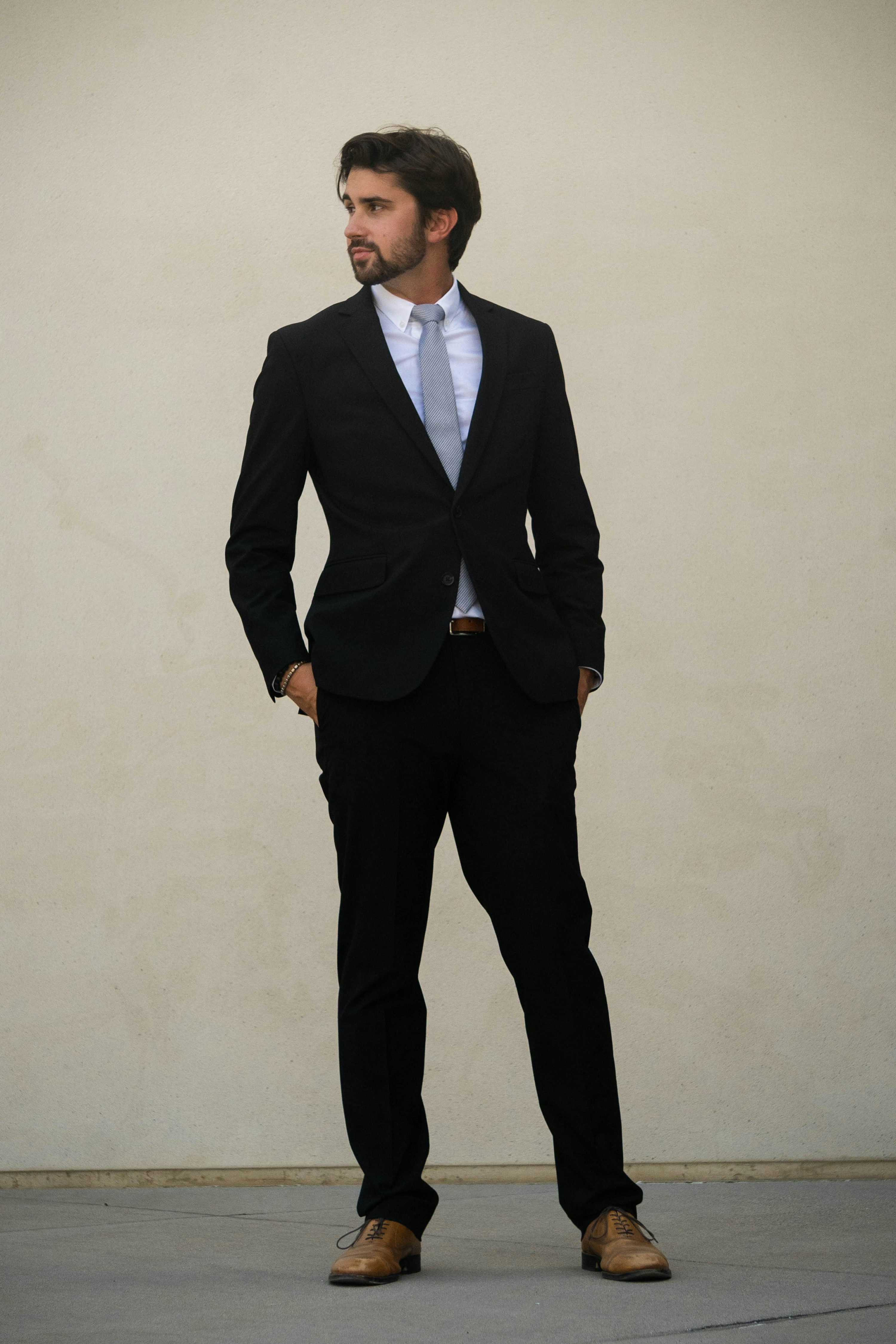 a fullbody shot of a man in a dark suit.  he's standing against a light beige wall, hands in pockets, looking off to the side with a somewhat serious expression. he's wearing a light blue shirt and a dark gray tie. his shoes are light brown oxfords. the overall style is professional and polished.
