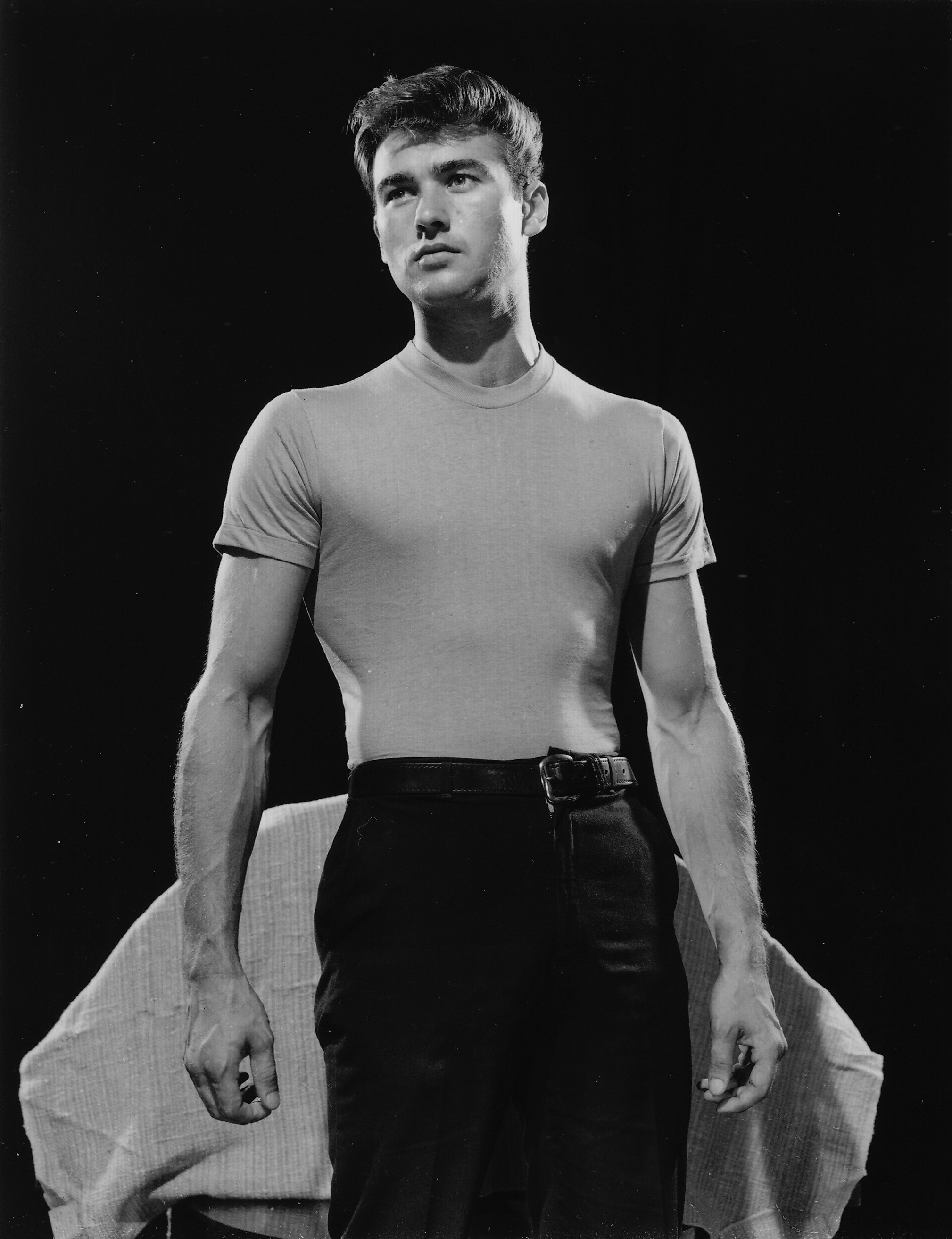 a black and white portrait of a young man, likely an actor, posed against a dark background. he wears a tightfitting, lightcolored tshirt that accentuates his physique and dark pants held with a belt. his hair is styled in a classic 1950s cut. he stands with his arms slightly away from his body, hands relaxed, and gaze directed upward. the lighting is dramatic, creating a strong contrast between the light and shadow, further emphasizing his muscular build. the overall feel is one of classic hollywood glamour and intensity.