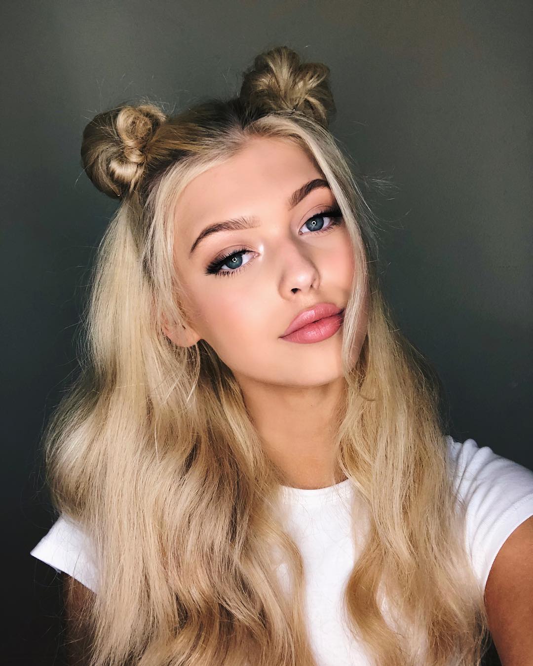 a portrait of a young woman with blonde hair styled in space buns. she has blue eyes and is wearing makeup, including eyeliner and lipstick. she is wearing a white shirt. the image appears to be a selfie.