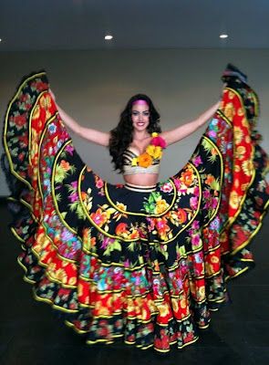 a woman in a colorful floral flamenco dress stands with arms outstretched. the dress is a multilayered, ruffled skirt with vibrant floral patterns in black, red, yellow, and green. she is wearing a matching crop top adorned with flowers, a pink headband, and has dark curly hair. the background is a plain wall with overhead lighting.