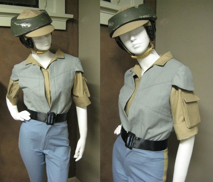 this is an image of two female mannequins dressed in matching spacethemed uniforms, both wearing helmets with attached goggles. the mannequins are positioned back to back, and both are facing forward, their heads turned to the side, to view their  profile. the mannequins are both dressed in grey jumpsuits with tan collars and sleeve accents, with black belts. they are both wearing spacethemed helmets. the one on the left appears to be a pilot, with a visor, and the one on the right appears to be a ground trooper, with a helmet and  goggles that are attached.  the background is a light, woodpaneled wall, with white trims.