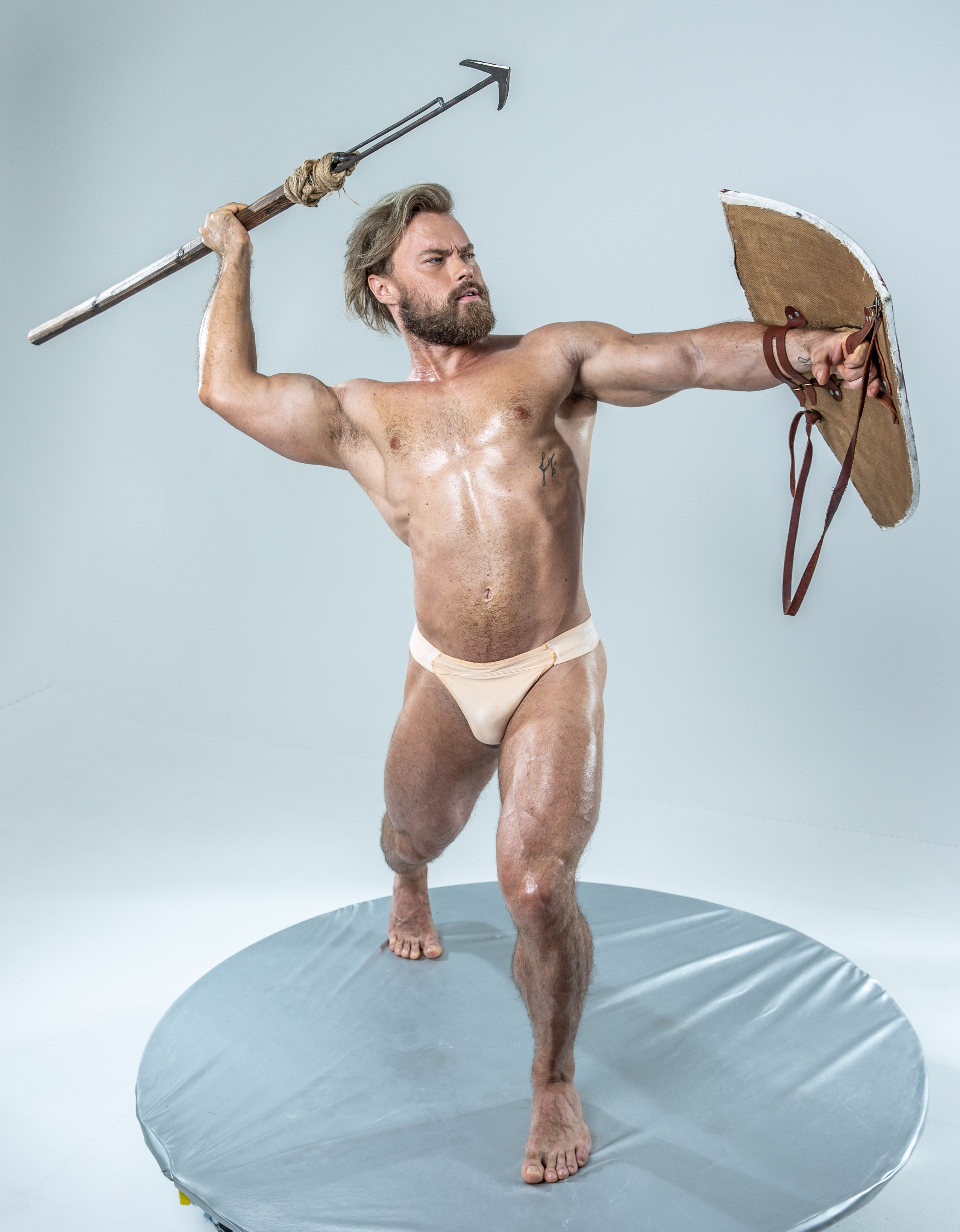 a muscular man, with sweat glistening on his tanned skin, stands poised for battle on a circular platform. he wields a spear in his right hand, held aloft in a dynamic pose, and a battered, round shield in his left. his intense gaze and powerful physique convey a sense of strength and readiness.  he is clad only in a simple loincloth, emphasizing the physique developed for combat.  the neutral background and soft lighting focus attention on the warrior's form and the textures of his weapons.