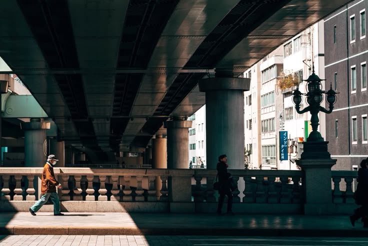 a photograph depicting a scene under a city overpass. two individuals are walking in opposite directions. the older man on the left is wearing a face mask, while the woman on the right is dressed in darker attire. the architecture is modern, with tall buildings and a large concrete overpass casting shadows. the setting appears to be a city in japan, judging by the building style and signage visible. the photograph is welllit with the sunlight highlighting the contrasts of light and shadow. the scene is serene and captures a moment in urban life.
